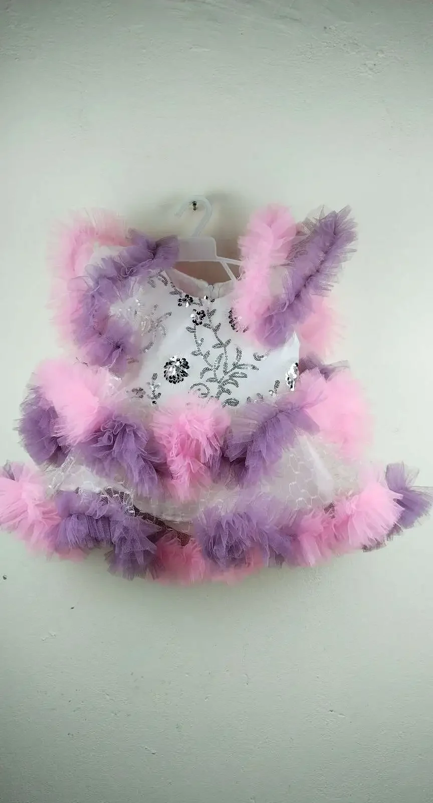 Pink & Purple Tulle Baby Dress with white bodice featuring silver floral embroidery and sequins, paired with a voluminous skirt made of layered white mesh and tulle, trimmed with alternating pink and purple ruffles, hanging on a white plastic hanger against a plain white wal