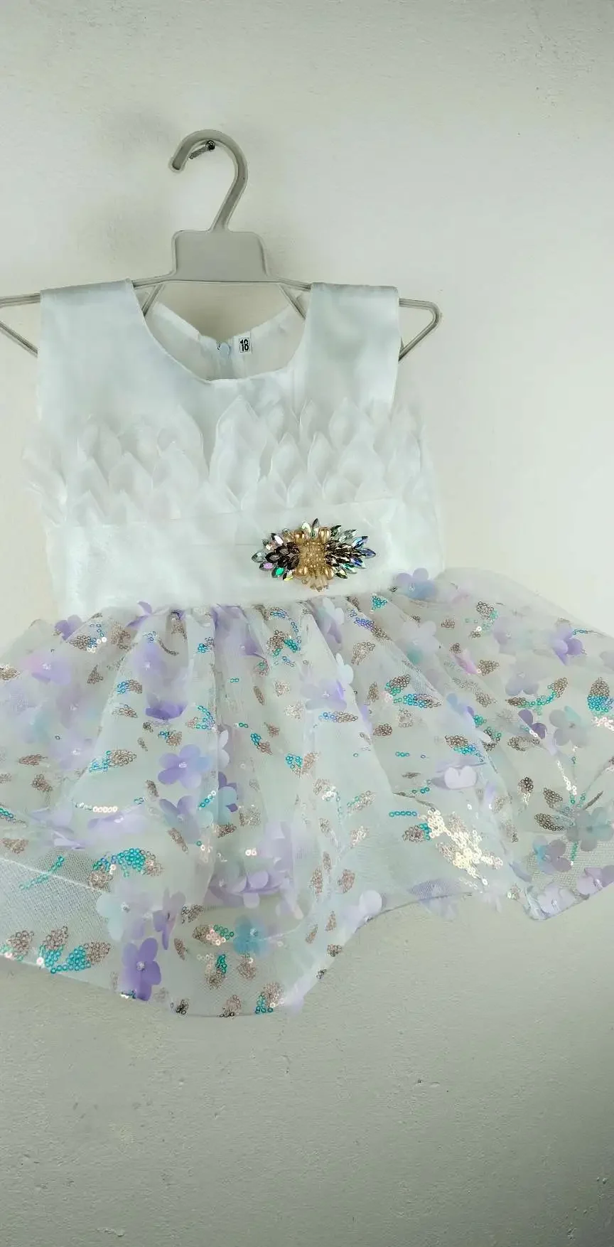 White sleeveless baby dress with textured leaf-patterned bodice, jeweled brooch at waist, and sheer tulle skirt adorned with pastel purple and blue flower appliqués and sequins, displayed on a hanger against a plain wall