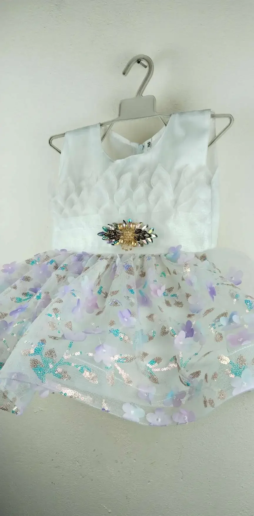 Elegant White Baby Dress with textured leaf-patterned bodice, jeweled brooch at waist, and sheer tulle skirt adorned with pastel purple and blue flower appliqués and sequins, displayed on a hanger against a plain wall