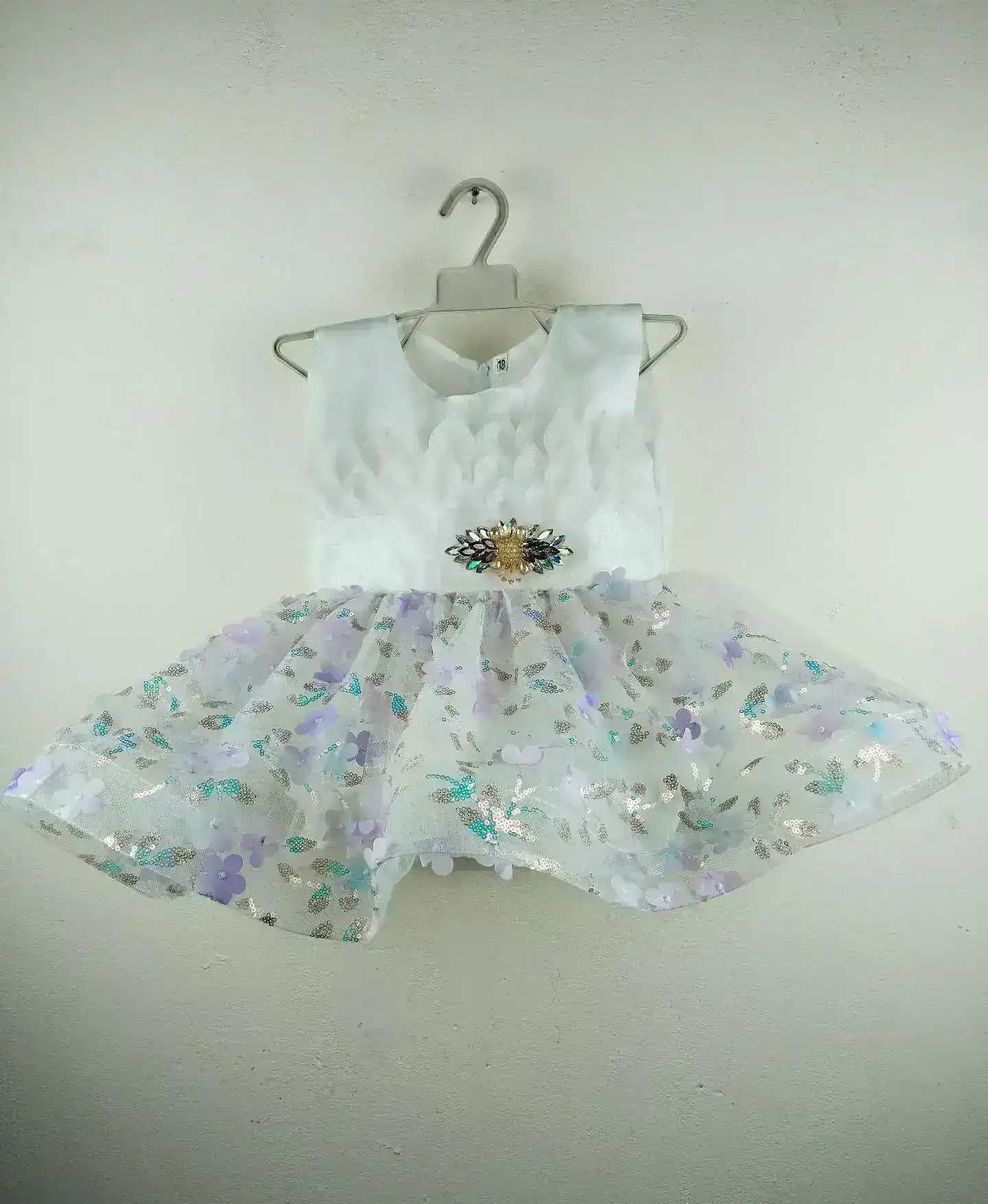 Elegant White Baby Dress with textured leaf-patterned bodice, jeweled brooch at waist, and sheer tulle skirt adorned with pastel purple and blue flower appliqués and sequins, displayed on a hanger against a plain wall