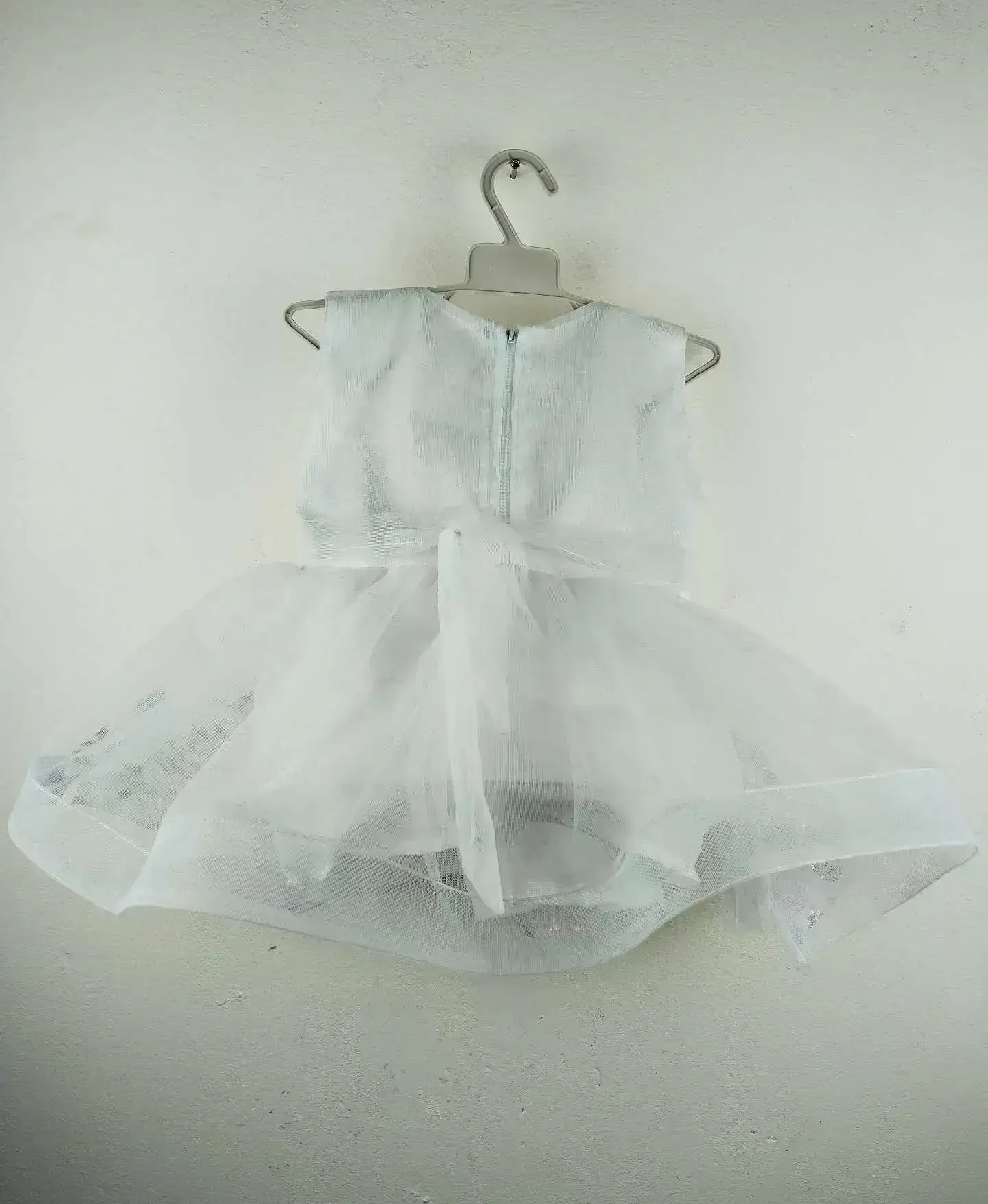 Elegant White Baby Dress with textured leaf-patterned bodice, jeweled brooch at waist, and sheer tulle skirt adorned with pastel purple and blue flower appliqués and sequins, displayed on a hanger against a plain wall