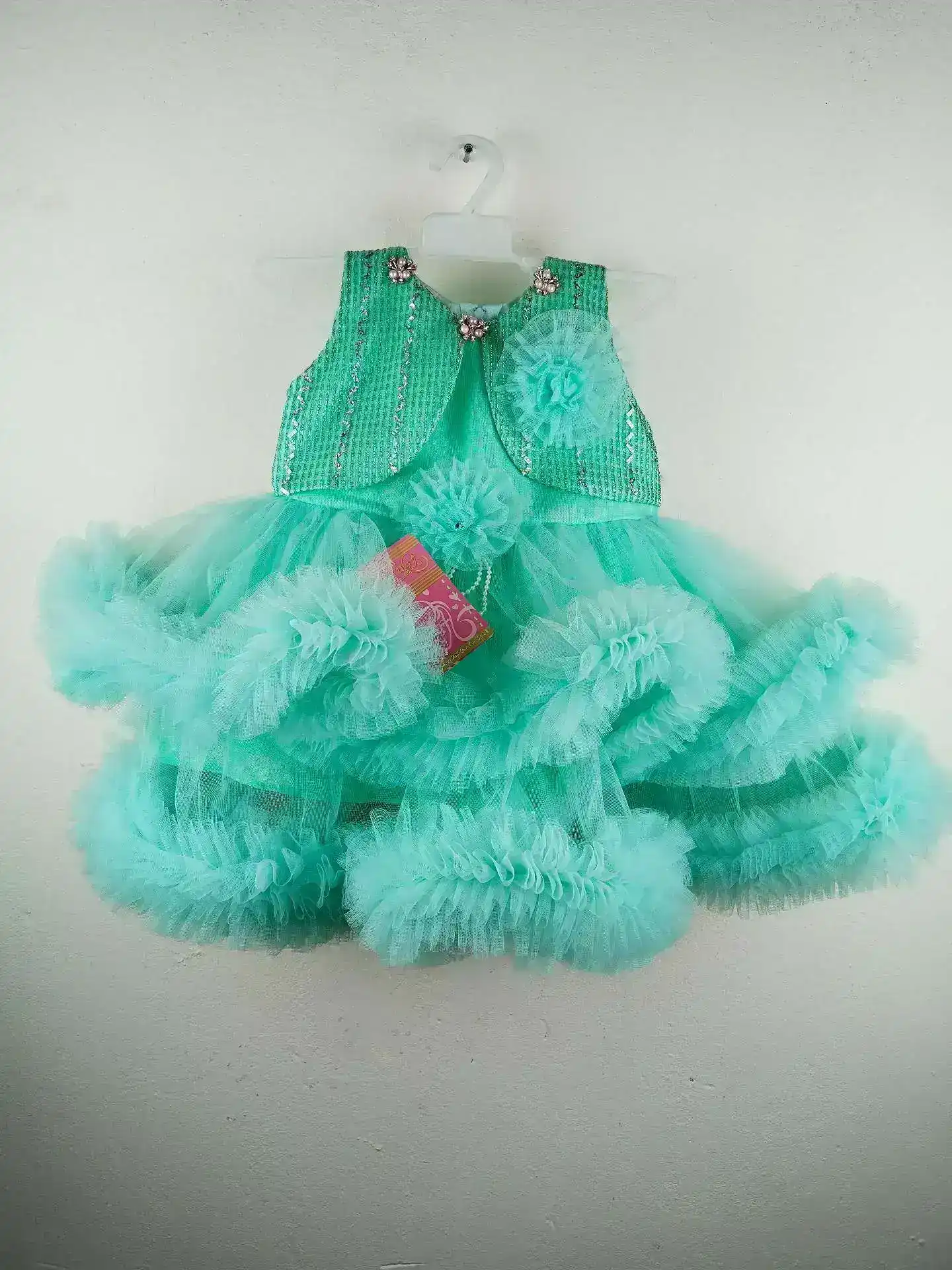 Mint Green Floral Baby Frock with Tulle Layers | GlamDusk with glittery vertical stripes, floral shoulder accents, and a voluminous layered tulle skirt adorned with ruffled details and tulle flowers, hanging on a white plastic hanger against a plain white wall