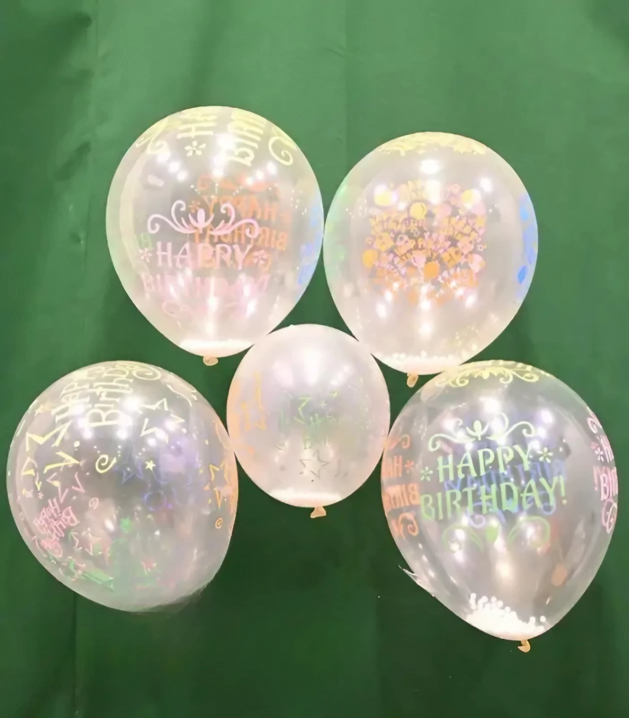 Transparent birthday balloons with colorful 'Happy Birthday' prints and small white confetti inside, displayed against a dark green background